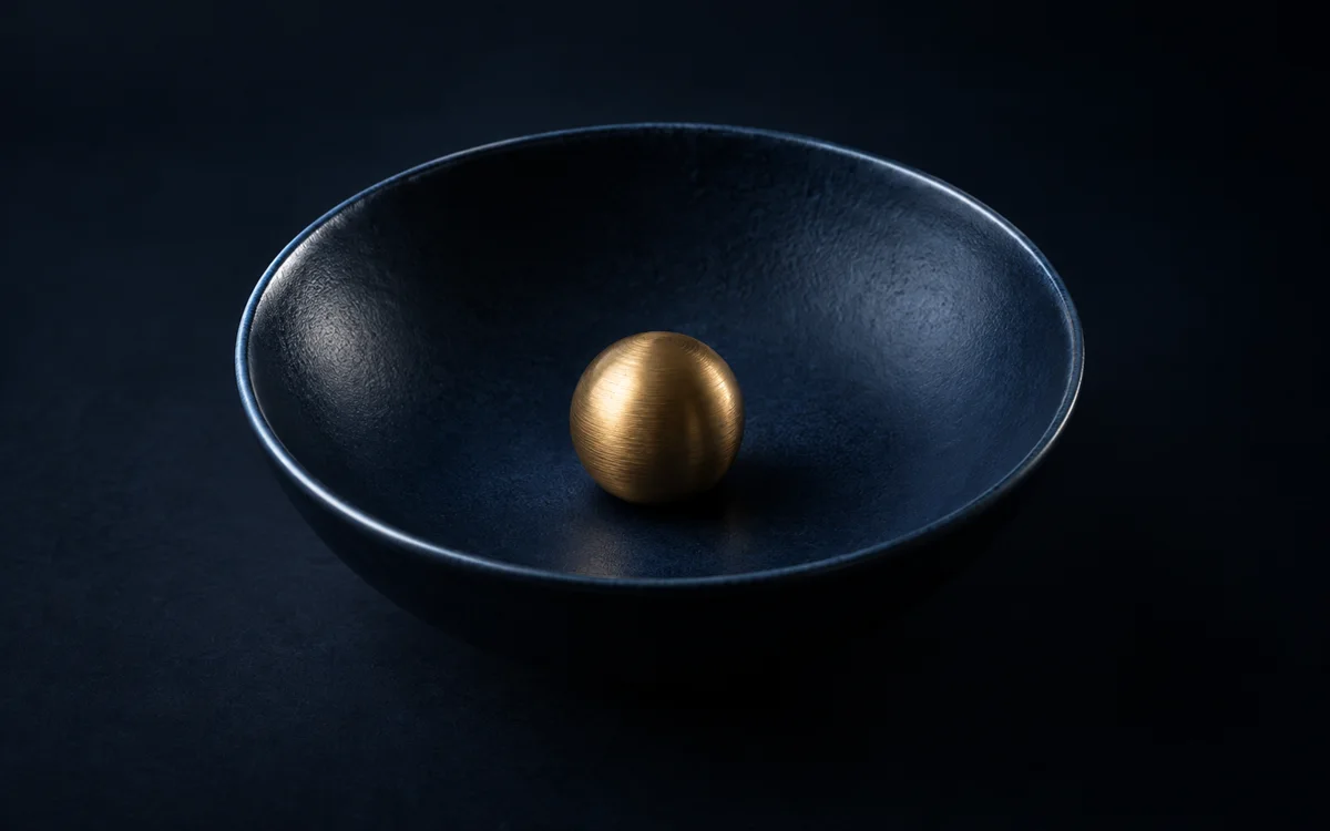 A brushed-brass sphere resting in a shallow deep-navy ceramic bowl